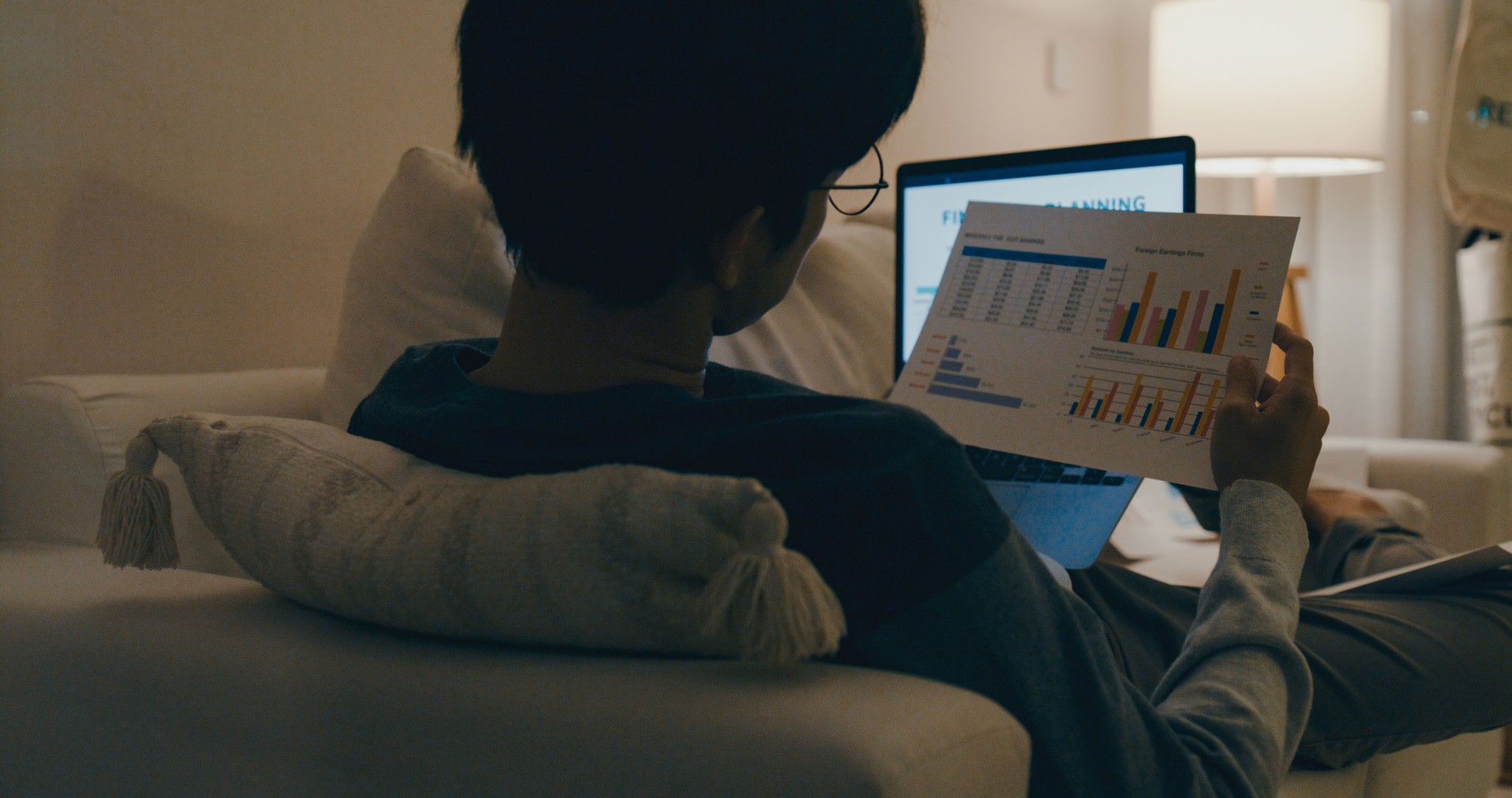 Young Asian man with eyeglasses lying on sofa, analyzing financial documents and working on laptop at night, showcasing dedication to work. Remotely and finance.